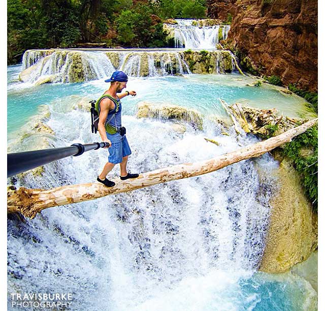 Man walking across a thin tree trunk over a plunging waterfall